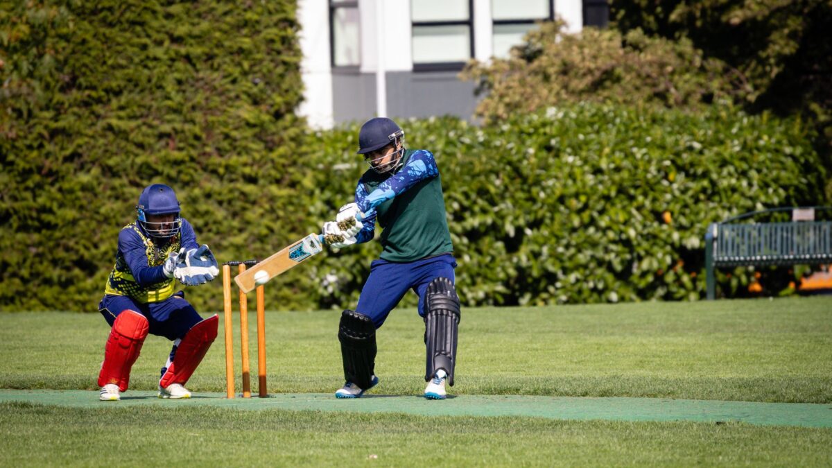Un batteur en tenue bleue et casque se tient prêt à frapper la balle devant les guichets, un gardien en tenue bleue et jaune derrière lui, sur une pelouse verte de cricket.