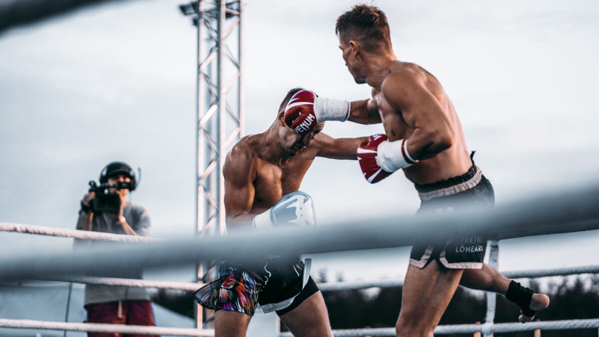 Deux boxeurs s'affrontent sur un ring en plein air. Celui de droite, gants rouges Venum et short noir, frappe son adversaire torse nu au visage. Un caméraman filme la scène au premier plan, sous un ciel gris et des projecteurs.