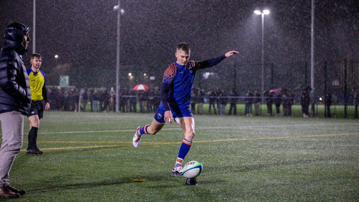 Un joueur en maillot bleu et orange frappe un ballon posé sur un tee sous la pluie, la nuit, sur un terrain synthétique éclairé par des projecteurs.