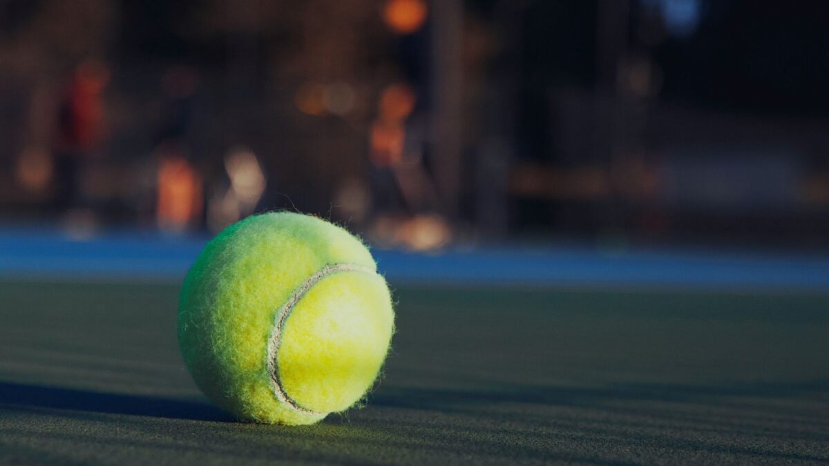 Une balle de tennis jaune fluo posée sur un court vert, en gros plan, avec un arrière-plan flou aux lumières orangées et un marquage bleu.