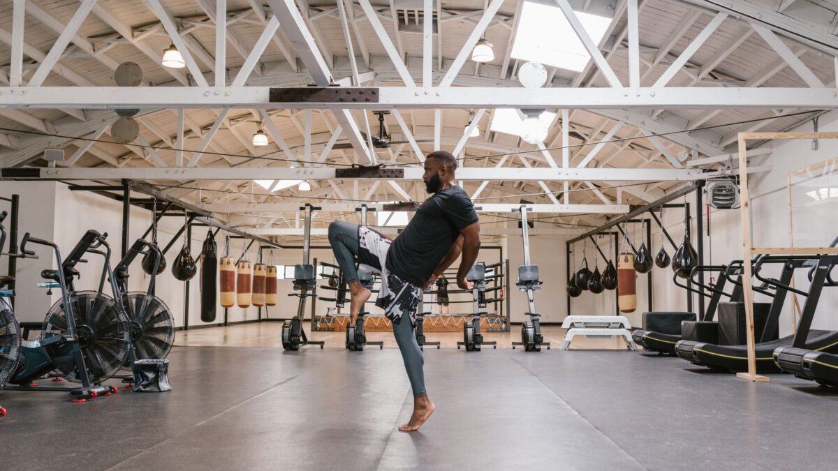 Un athlète de MMA s'exerce à l'équilibre sur une jambe dans une salle de sport avec des sacs de frappe et des machines de fitness.