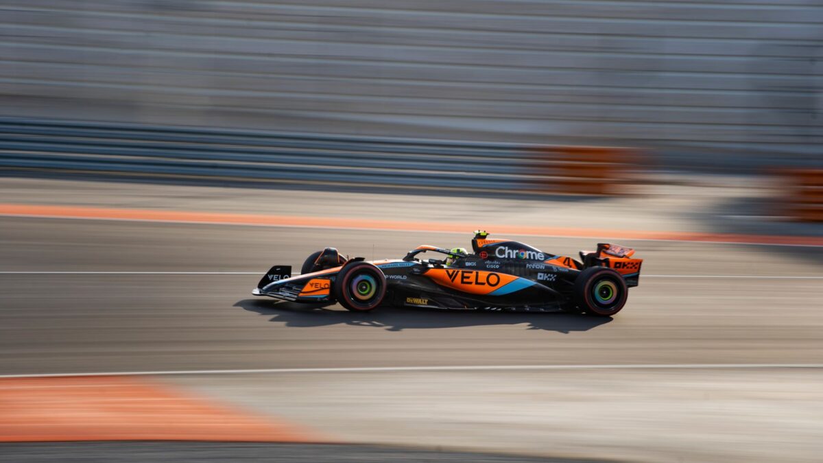 Une monoplace McLaren orange et noire, aux sponsors VELO, Chrome et DEWALT, roule à grande vitesse sur un circuit de Formule 1, avec un effet de filé sur le décor et des barrières orange en arrière-plan.