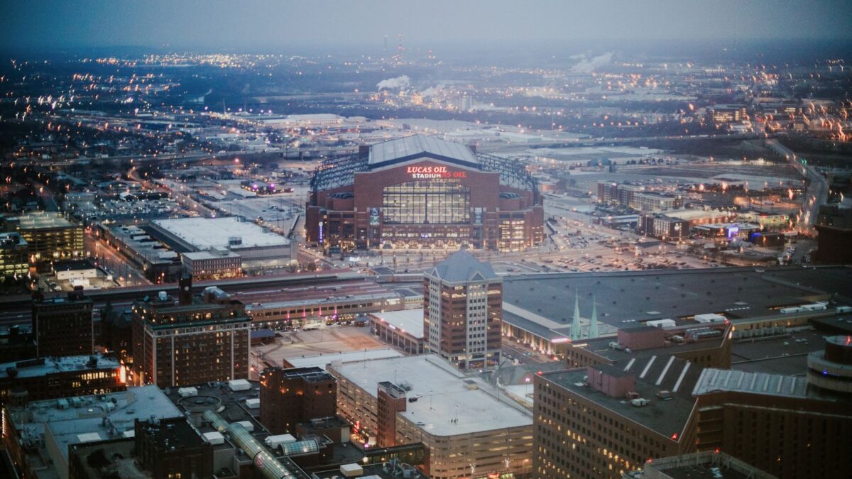 Vue aérienne d'une ville en soirée, avec le stade Lucas Oil illuminé au centre, entouré de bâtiments et de routes.
