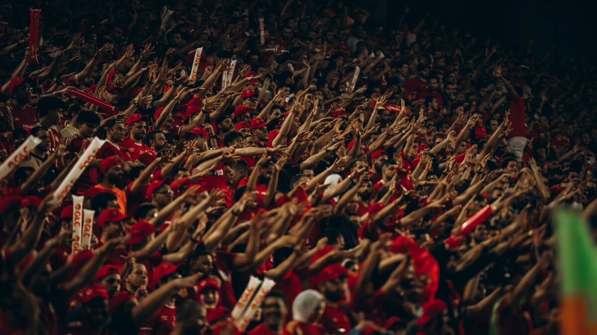 Foule de supporters en rouge, bras levés, tenant des objets lumineux dans un stade sombre.