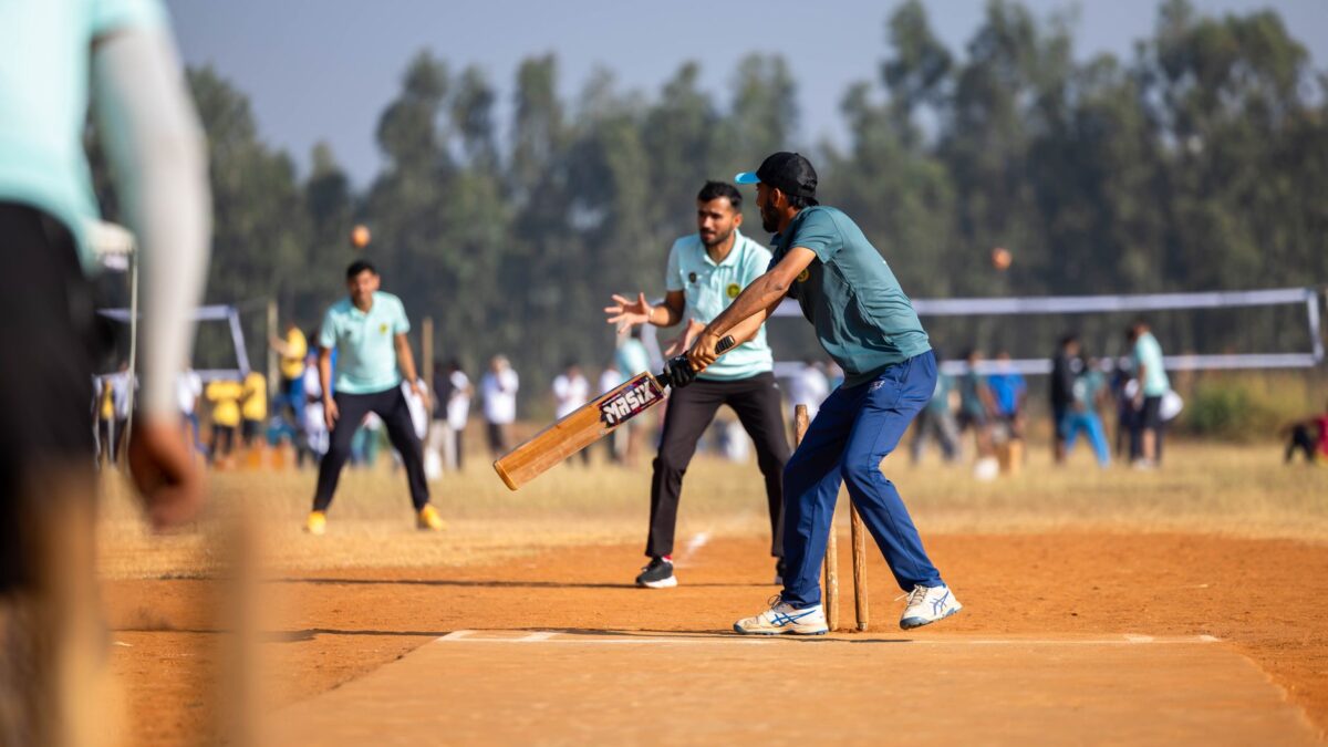 Un batteur en tenue bleue et casquette noire frappe une balle lors d'un match de cricket sur un terrain en terre battue, avec des joueurs en turquoise et des spectateurs en arrière-plan.