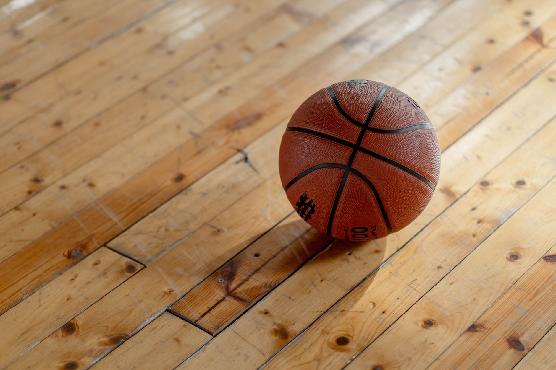 A basketball resting on a worn wooden plank floor in warm light, with a soft shadow beside it.