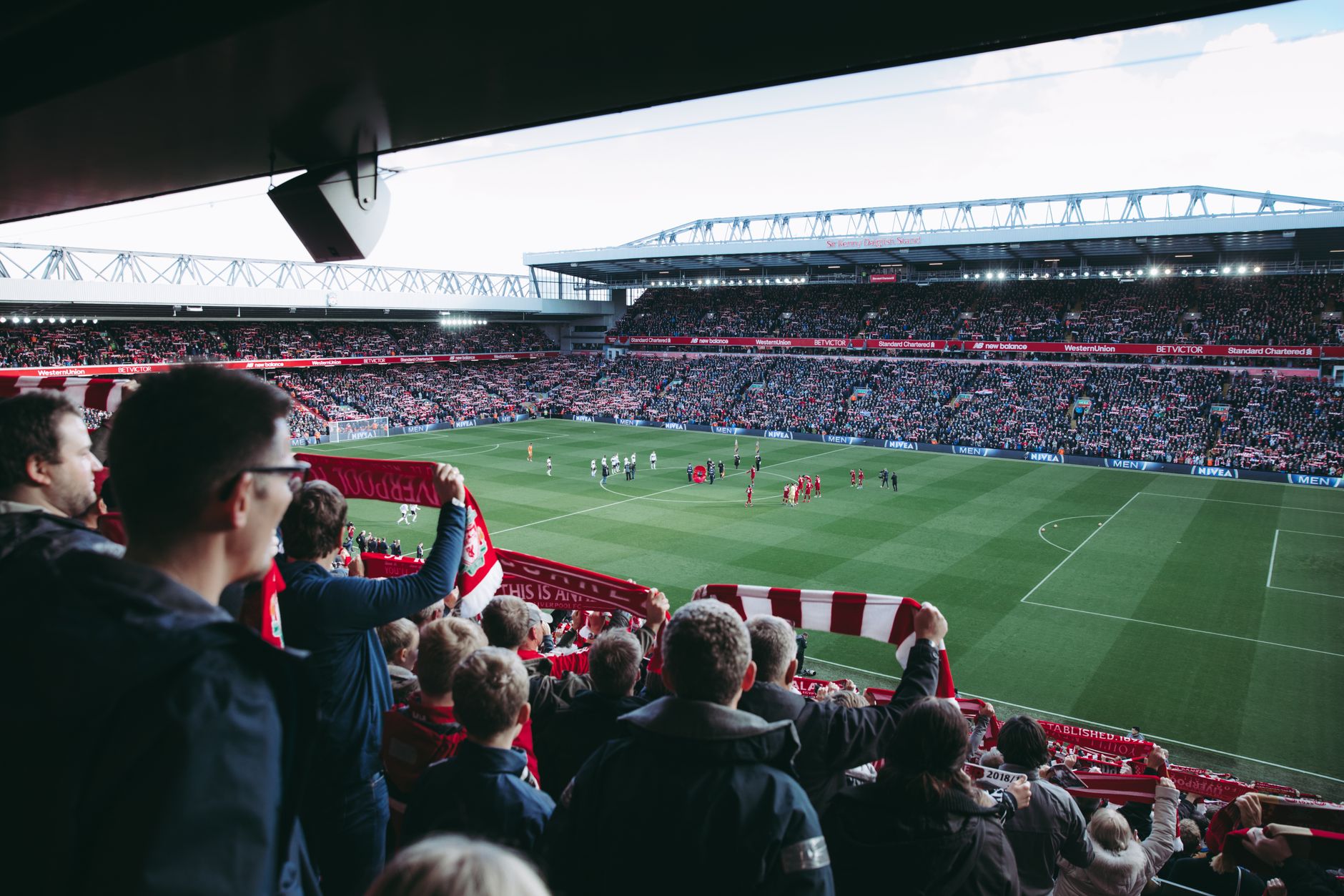 Supporters de dos levant des écharpes rouges dans une tribune, face à un stade de football plein et à des joueurs sur la pelouse.