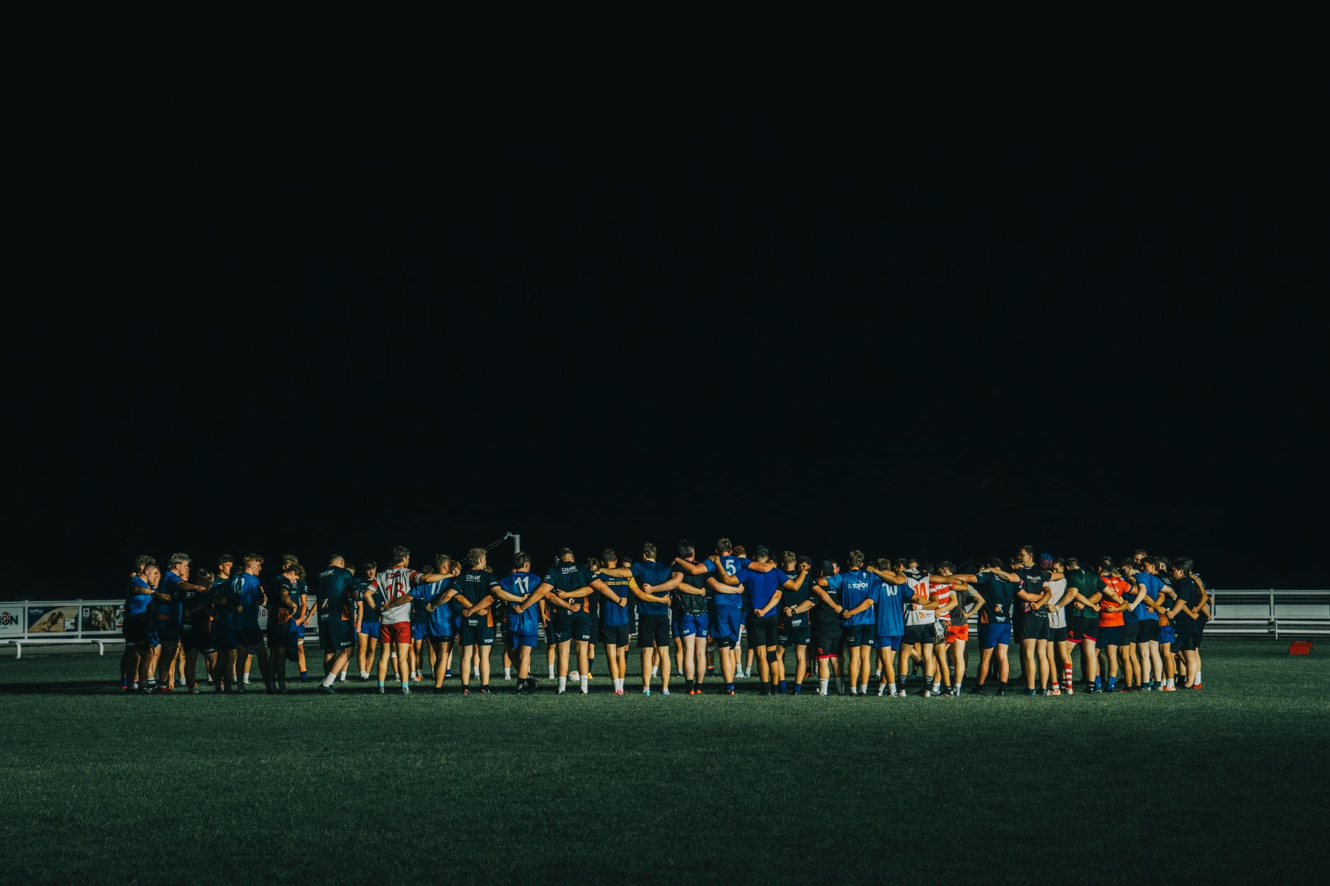 Team players in a wide huddle with arms around shoulders on a grass field at night under a dark sky.